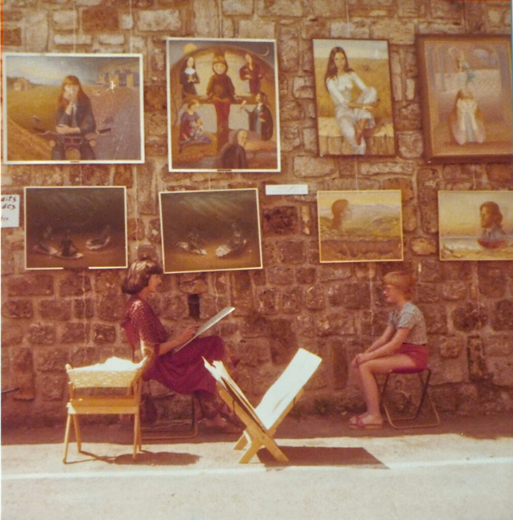 The artist sketching a boy outdoors in front of a large wall on which many of her paintings are hanging.