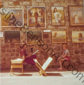 The artist sketching a boy outdoors in front of a large wall on which many of her paintings are hanging.